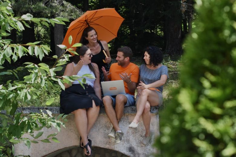 A diverse group of adults enjoying a conversation in nature, sitting with a laptop and umbrella on a sunny day.