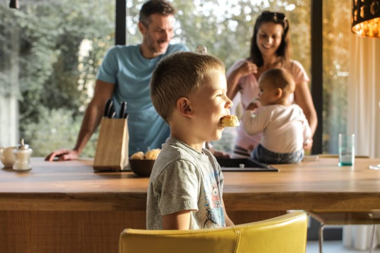 Side view of playful boy eating cookie while sitting at table with parents and sibling on background spending time at home